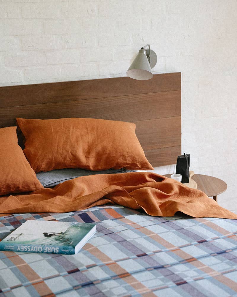 Bed with orange bedding and plaid blanket, wooden headboard, and lamp on a white brick wall background.