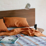 Bed with orange bedding and plaid blanket, wooden headboard, and lamp on a white brick wall background.