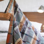 Person holding a plaid blanket over a bed with a wooden headboard.