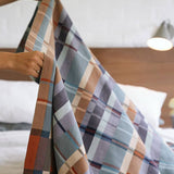 Person holding a plaid blanket over a bed with a wooden headboard.