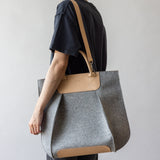 A person in black attire holds the versatile Frankie Tote by graf lantz—an elegantly structured gray bag with tan leather straps and accents. Set against a simple, light gray background, this tote blends style with functionality.