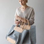 A person with short blond hair in a beige sweater holds the large Frankie Tote by graf lantz, featuring gray and tan hues with leather handles. The simple gray background adds a minimalist touch, enhancing the stylish look.