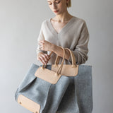 A person with short blond hair in a beige sweater holds the large Frankie Tote by graf lantz, featuring gray and tan hues with leather handles. The simple gray background adds a minimalist touch, enhancing the stylish look.