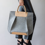 A person carries a large gray and beige Frankie Tote by graf lantz, crafted from Merino Wool, complementing their black leather pants and heeled ankle boots against a plain light gray backdrop.