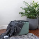 Green storage basket with a gray blanket next to a potted plant on a wooden floor.
