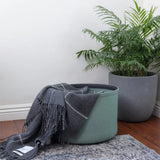 Green storage basket with a gray blanket next to a potted plant on a wooden floor.