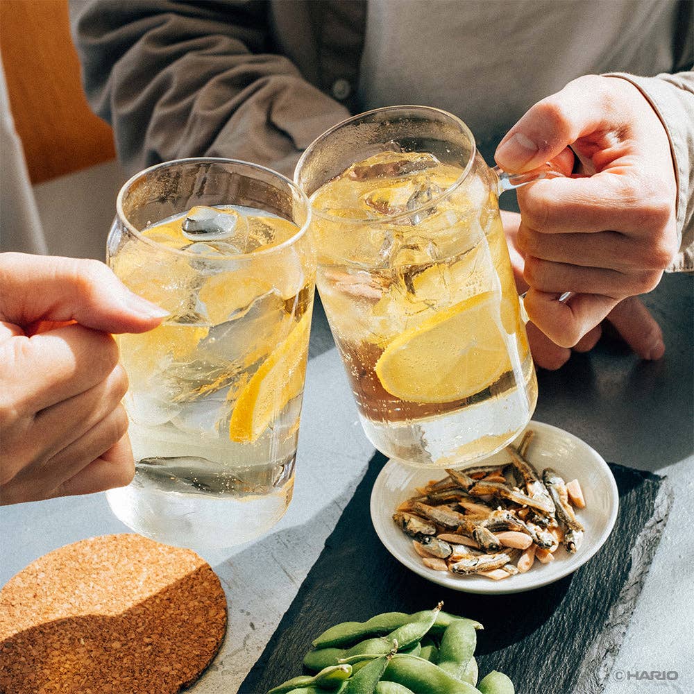 Two glasses of lemonade with ice being held by hands, with a small plate of snacks on a table.