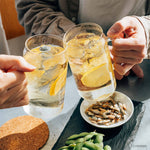 Two glasses of lemonade with ice being held by hands, with a small plate of snacks on a table.