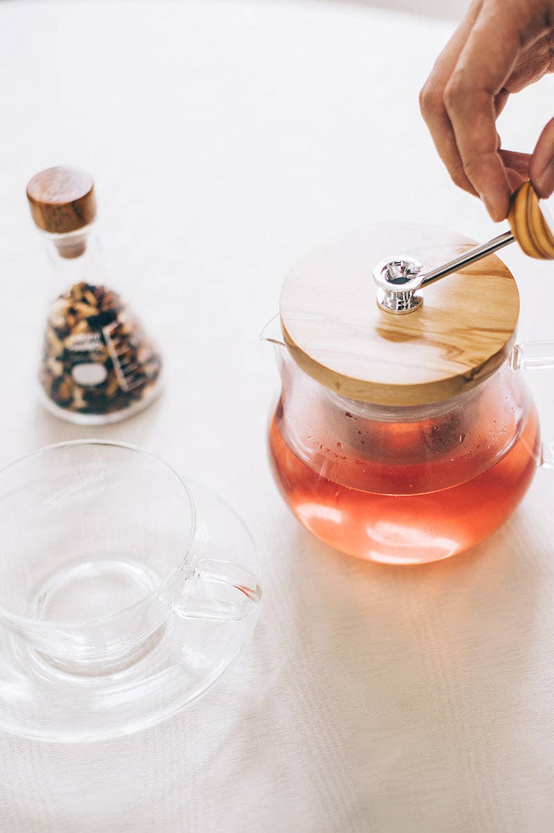Glass teapot with wooden lid on a white surface, with a hand holding the tea strainer handle
