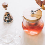 Glass teapot with wooden lid on a white surface, with a hand holding the tea strainer handle