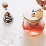 Glass teapot with wooden lid on a white surface, with a hand holding the tea strainer handle