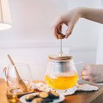 Person making tea with a teapot on a wooden table.