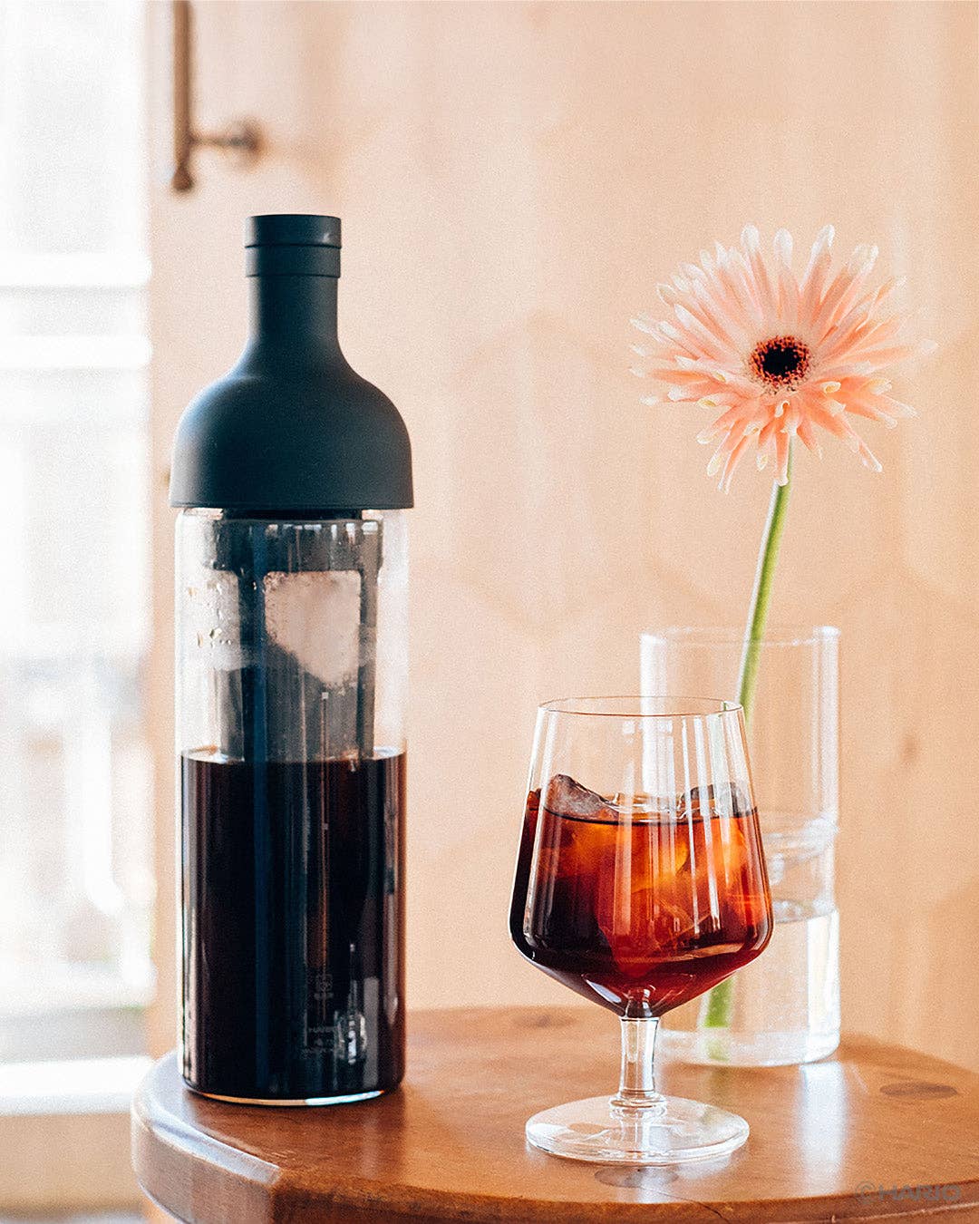 Bottle of dark liquid next to a glass with a floral arrangement on a wooden surface.
