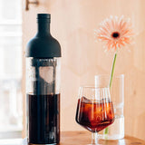 Bottle of dark liquid next to a glass with a floral arrangement on a wooden surface.