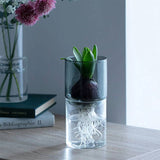 Clear glass vase with a plant, books, and flowers on a wooden surface.