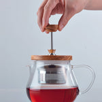 Hand pulling up metal tea strainer of glass teapot with wooden lid filled with tea, on a light gray background