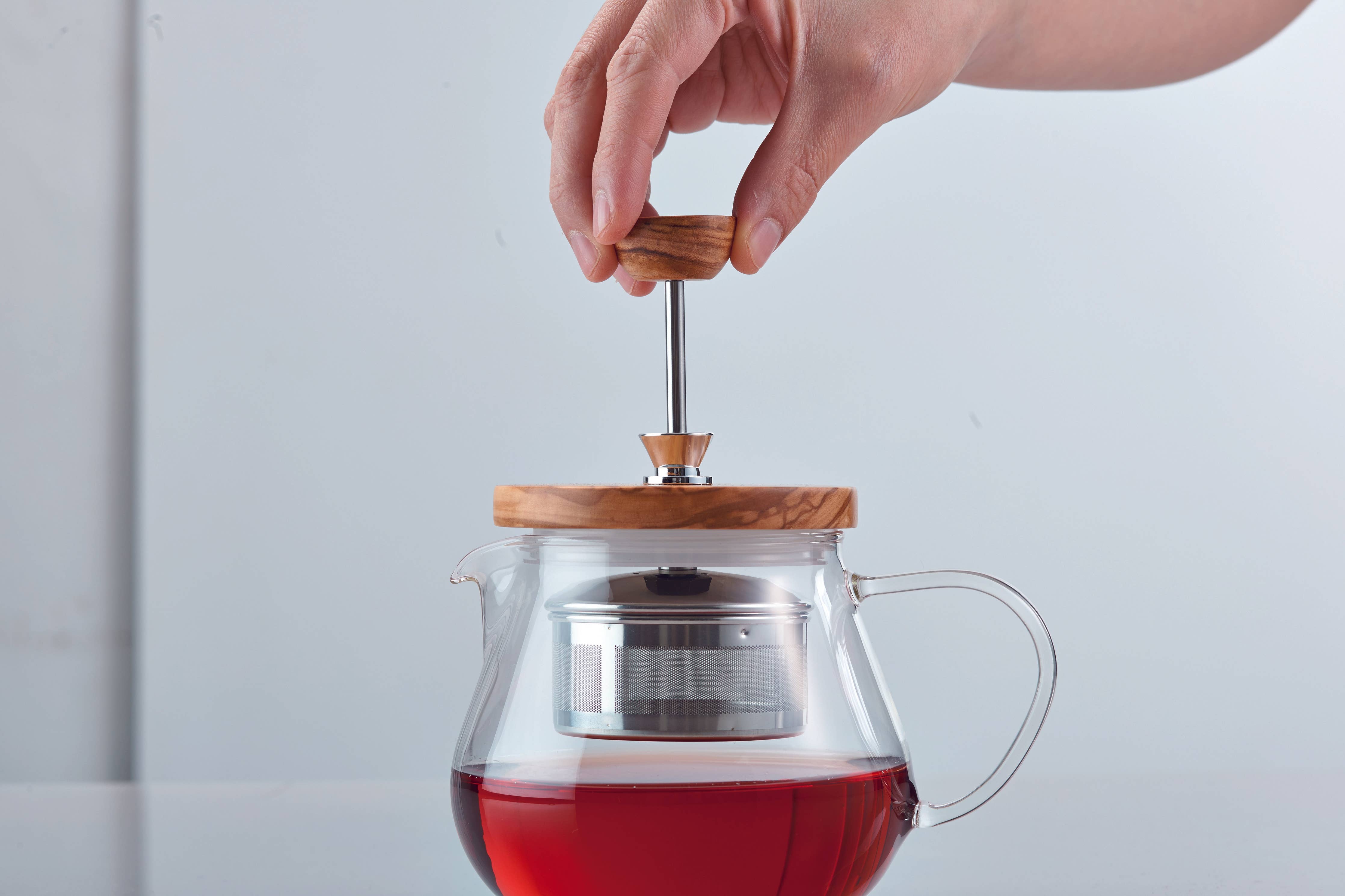 Hand pulling up metal tea strainer of glass teapot with wooden lid filled with tea, on a light gray background