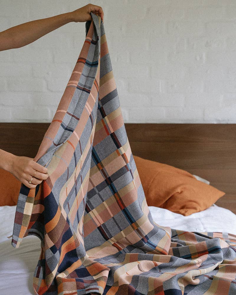 Person holding a plaid blanket over a bed with orange pillows.
