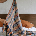 Person holding a plaid blanket over a bed with orange pillows.