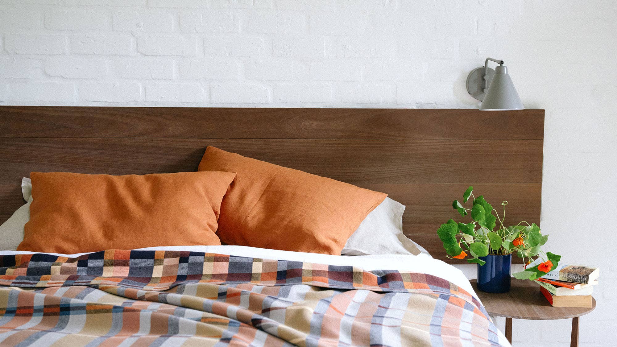 Bed with orange pillows and a plaid blanket against a white brick wall.