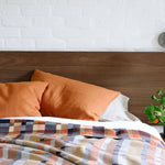 Bed with orange pillows and a plaid blanket against a white brick wall.