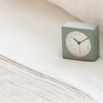 Square light green clock on a bed with white bedding