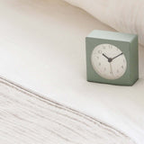Square light green clock on a bed with white bedding