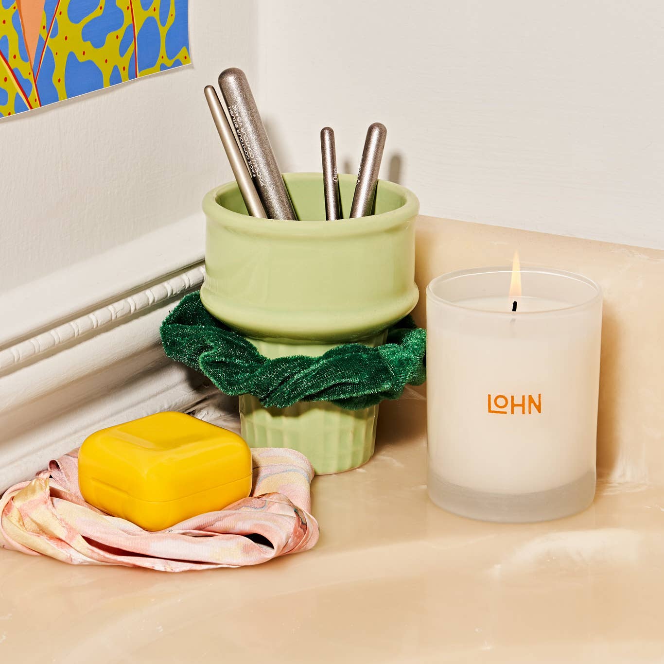Candle with 'LOHN' branding, green container with pens, yellow soap, and pink towel on a surface.