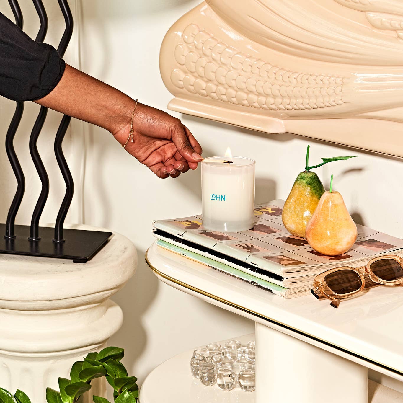 Hand lighting a Lohn candle next to a magazine, sunglasses, and pears on a table.