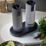 Two pepper grinders on a black tray with herbs and spices in the background