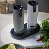 Two pepper grinders on a black tray with herbs and spices in the background