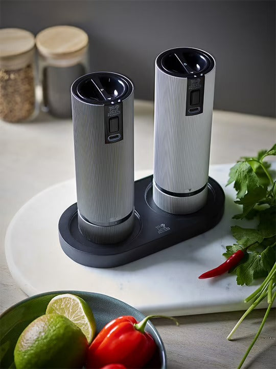 Two pepper grinders on a black tray with herbs and spices in the background
