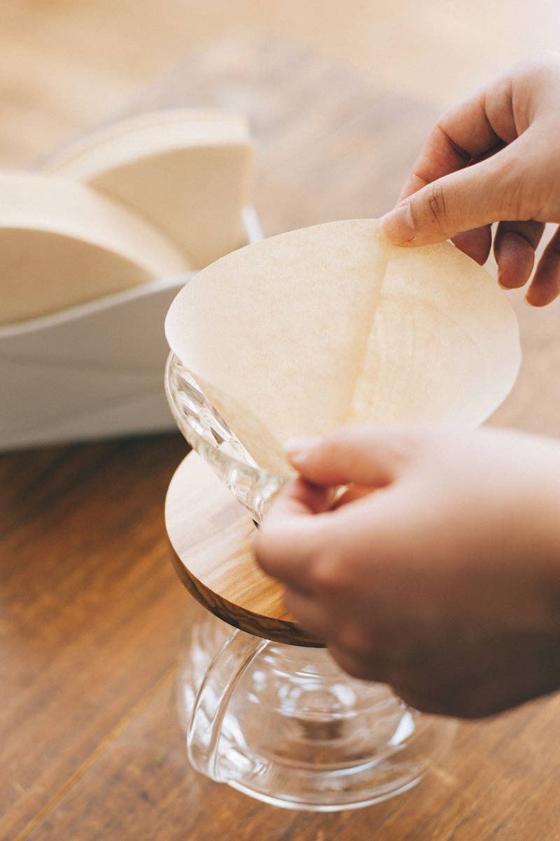 Person holding a paper coffee filter over a glass coffee dripper and carafe on a wood table