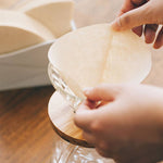 Person holding a paper coffee filter over a glass coffee dripper and carafe on a wood table