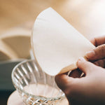 Person holding a natural paper filter over a clear coffee dripper 