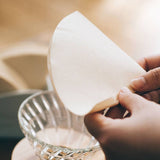 Person holding a natural paper filter over a clear coffee dripper 