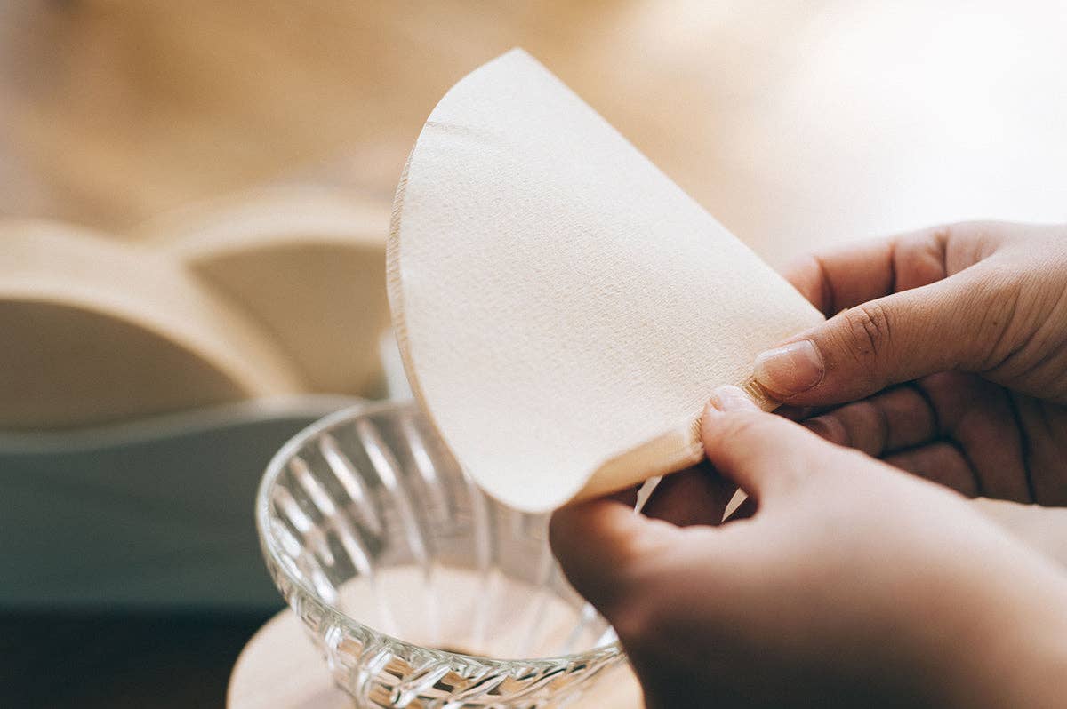 Person holding a natural paper filter over a clear coffee dripper 