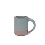 A medium sized ceramic mug with handle in a blue-grey color featuring iron speckles and unglazed rim.