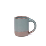 A medium sized ceramic mug with handle in a blue-grey color featuring iron speckles and unglazed rim.