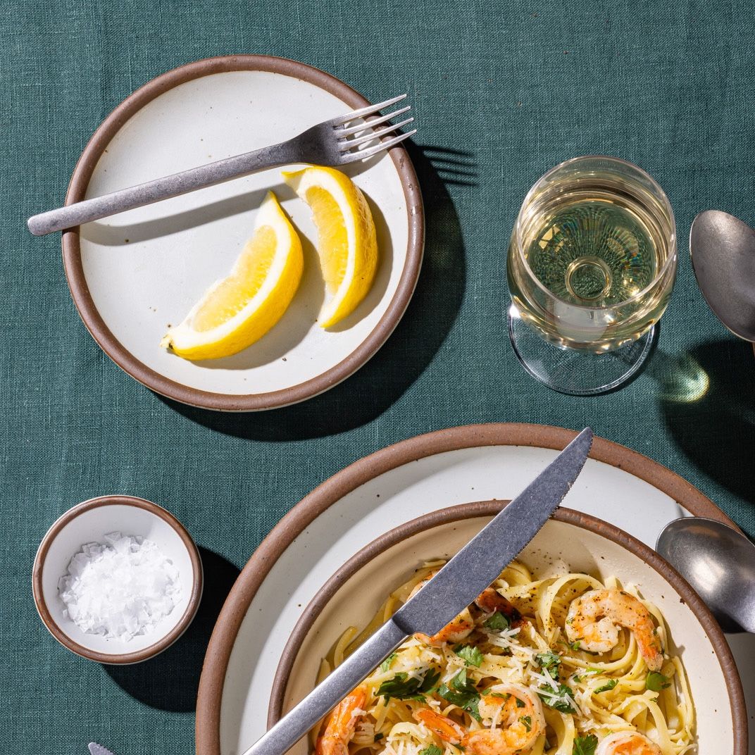 A ceramic cake plate with 2 lemon slices on it and a silver fork, also nearby is a place setting with pasta and a wine glass.