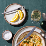 A ceramic cake plate with 2 lemon slices on it and a silver fork, also nearby is a place setting with pasta and a wine glass.