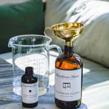 Large bottle with funnel, small bottle, and measuring cup on a marble surface with a couch in the background.