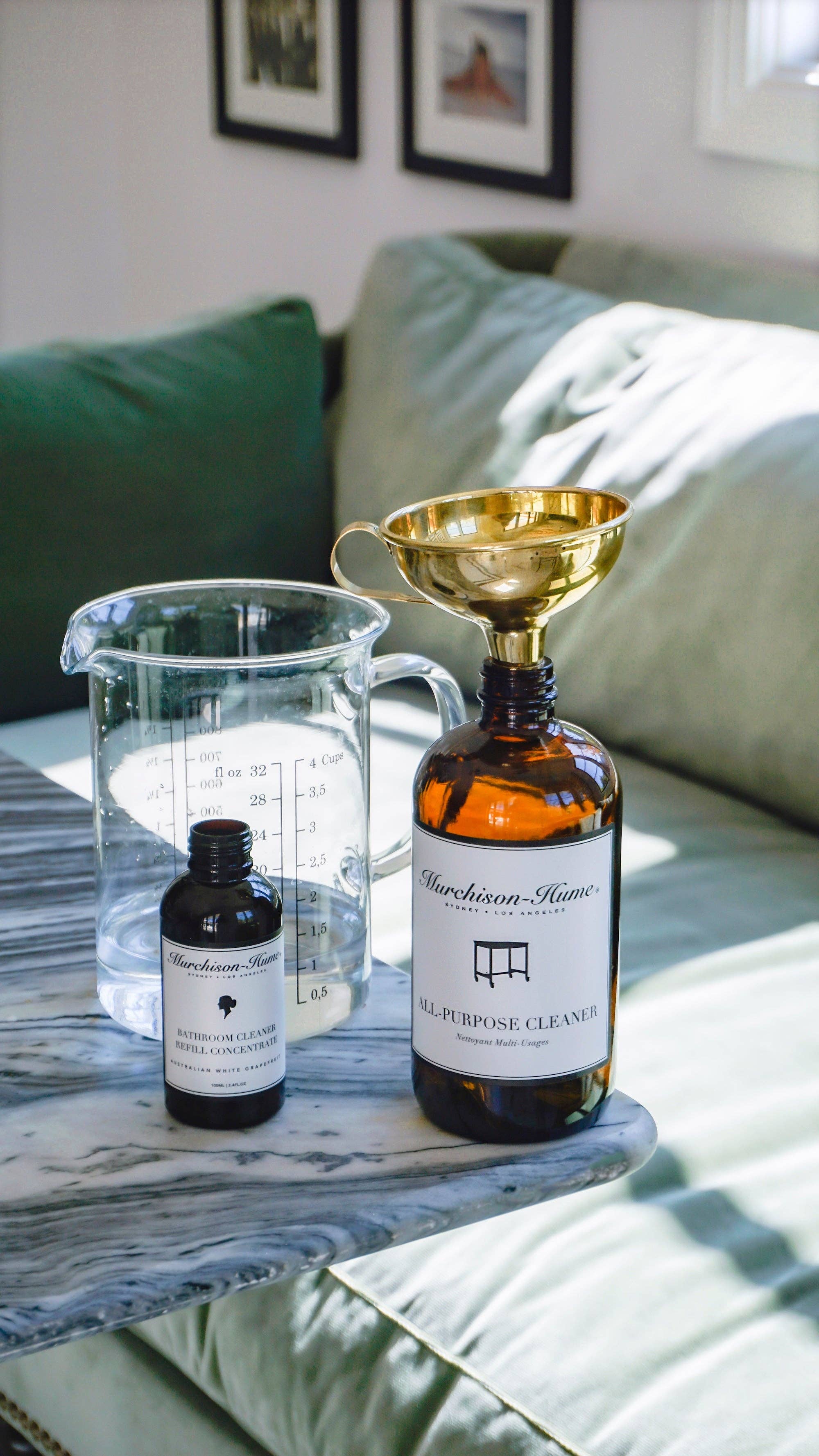 Large bottle with funnel, small bottle, and measuring cup on a marble surface with a couch in the background.