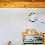 Brown clock mounted on white wall above wooden shelf with books and decorative items.