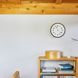 Brown clock mounted on white wall above wooden shelf with books and decorative items.