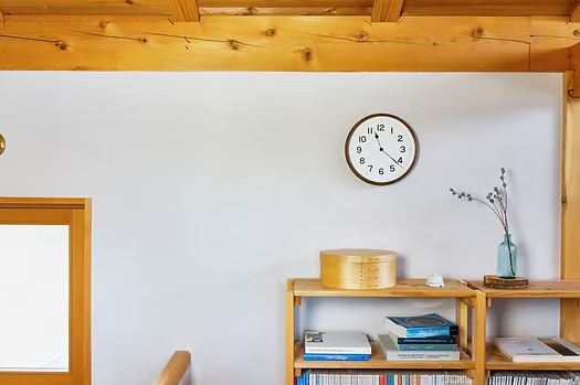 Brown clock mounted on white wall above wooden shelf with books and decorative items.