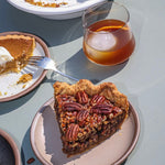 Slice of pecan pie on a warm pale brown ceramic dessert plate beside a glass and a partially eaten pumpkin pie slice.