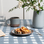 On a blue crosshatch tablecloth sits a cake plate in a blue-grey color with toast and spoon, nearby is a small mug and vase.
