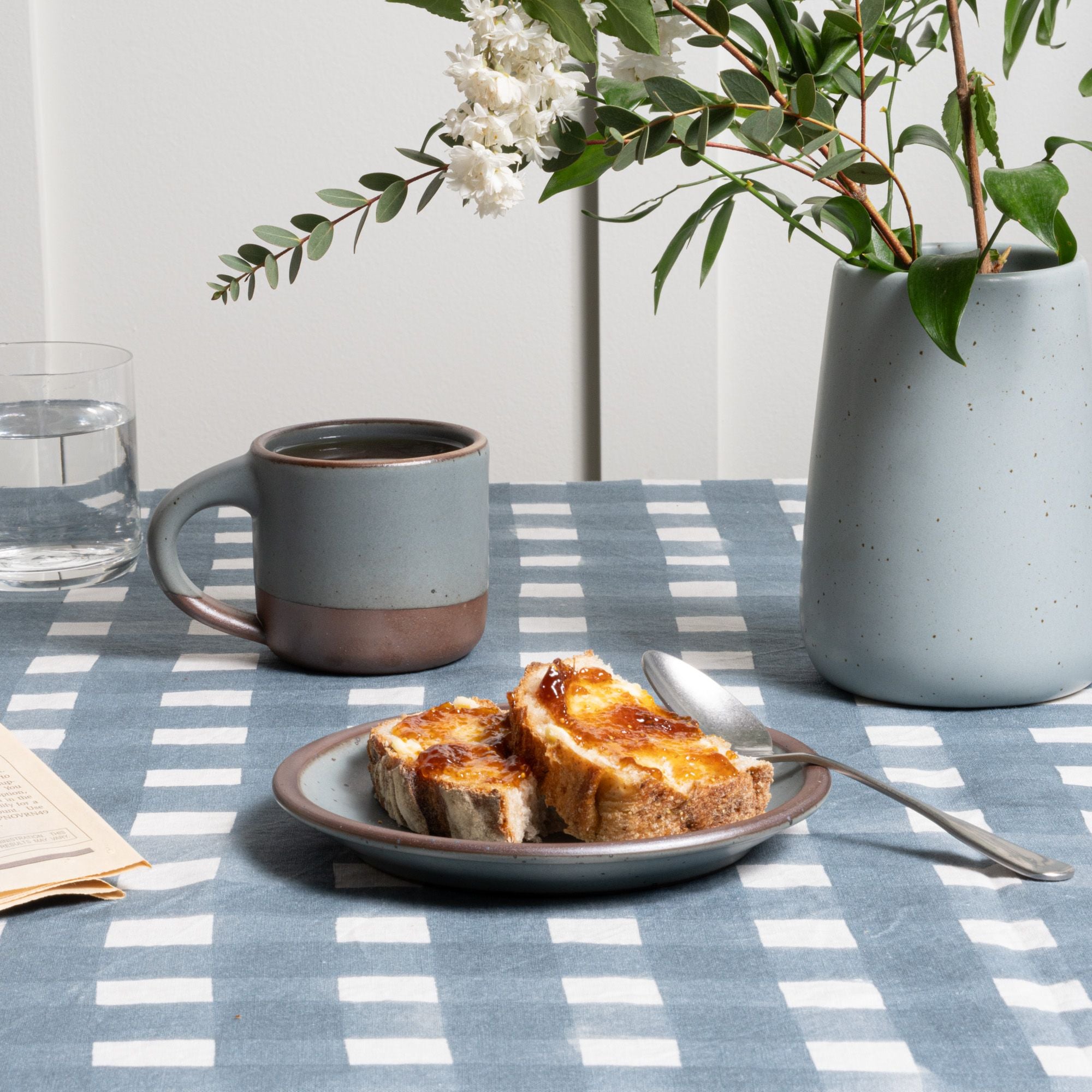 On a blue crosshatch tablecloth sits a cake plate in a blue-grey color with toast and spoon, nearby is a small mug and vase.
