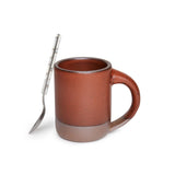 A spoon leaning on a medium sized ceramic mug with handle in a cool burnt terracotta color featuring iron speckles and unglazed rim and bottom base.